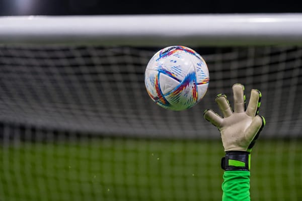 A dramatic overhead close-up captures a white soccer ball frozen in midair as it slips just beyond the fingertips of a lone goalkeeper's outstretched glove. Only the glove is visible, emphasi